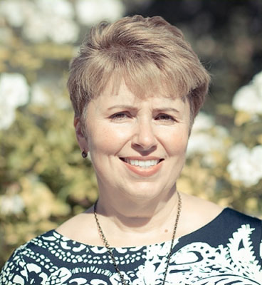 A smiling woman with short hair, wearing a dark top with a patterned sleeve, poses for a portrait outdoors.