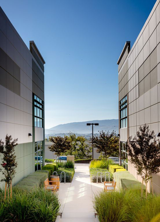 The image depicts a modern multi-story building with a walkway leading to an entrance, set against a clear sky with mountains visible in the distance.