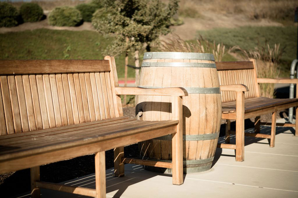 The image shows a wooden barrel-shaped planter placed on top of a park bench, with a background view of an outdoor seating area featuring more benches and natural surroundings.