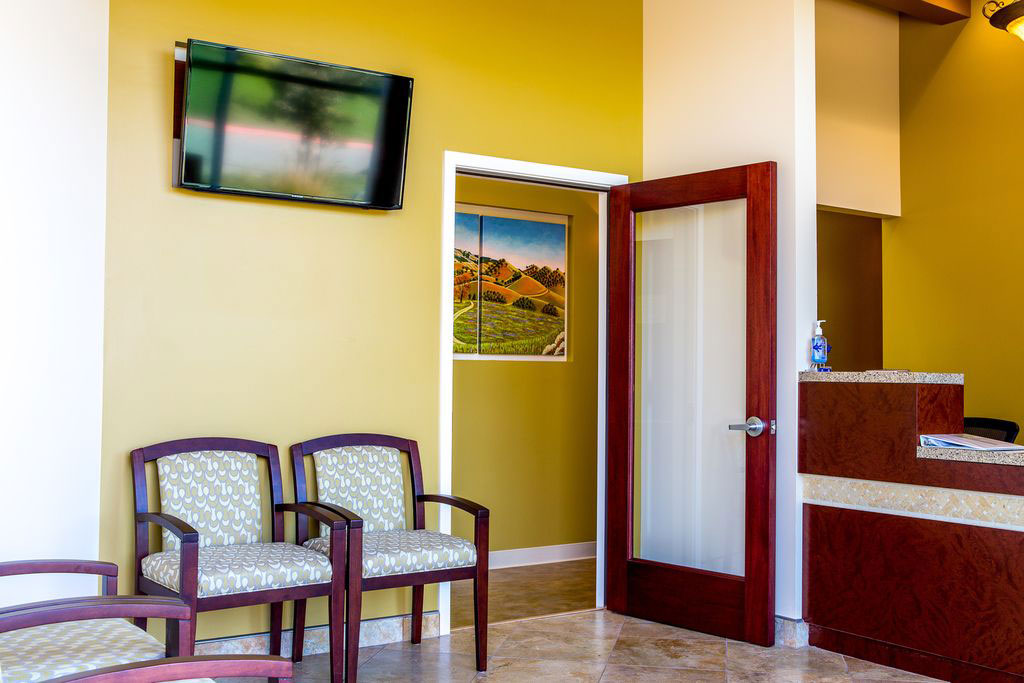 The image shows an interior space with a waiting area featuring chairs and a television screen on the wall, leading to a hallway with a door and a sign, which appears to be a medical office setting.