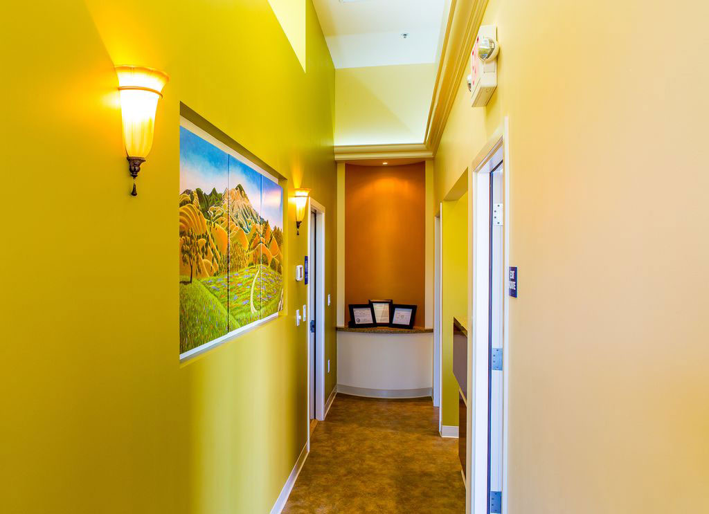 The image shows a well-lit hallway with yellow walls, a door on the right, and artwork on the wall.