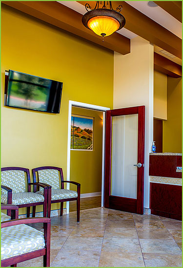 The image shows an interior view of a waiting room area with a color scheme of yellow walls, tan tiles, and brown trim, featuring a television mounted on the wall above a reception desk, and a seating area with chairs and a table.