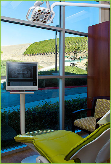 The image shows an interior view of a dental office with modern equipment, including a TV screen displaying dental images, a chair with a green cushion, and a window overlooking a street scene with a clear sky and hills in the background.