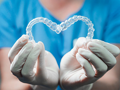The image shows a pair of clear braces held by gloved hands with a blue surgical gown background.