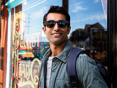 A young man wearing sunglasses, a backpack, and a jacket stands confidently in front of a storefront with a sign reading  Sweet Treats.