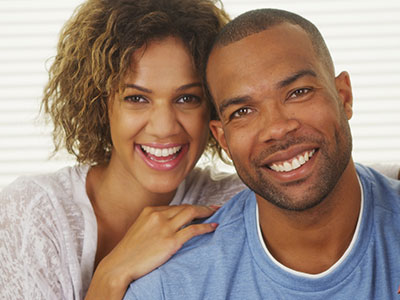 A man and woman smiling at each other with the man wearing a necklace and both standing close together.