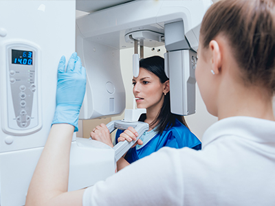 The image shows a woman standing in front of a large piece of medical equipment, which appears to be an MRI machine, with another person looking at her through a window.