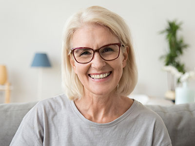 The image shows a woman with blonde hair, wearing glasses and a light-colored top, smiling at the camera against a blurred indoor background.