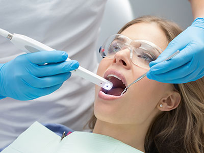 The image shows a dental professional using a device to clean teeth while a patient sits with their mouth open, wearing a blue surgical mask and protective eyewear.