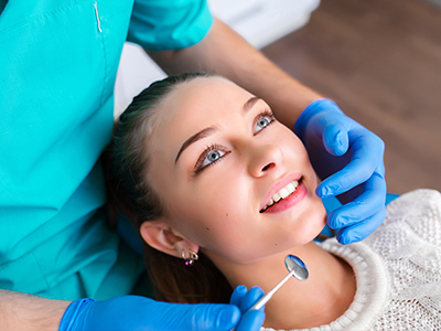 The image shows a female patient seated in a dental chair, receiving dental care from a professional wearing blue gloves and a face mask, with the professional using a dental instrument to examine her teeth while she smiles at the camera.