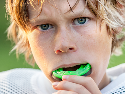 The image shows a young male athlete with blonde hair, wearing a football jersey, holding a green mouthguard in his mouth, looking directly at the camera with an intense expression.