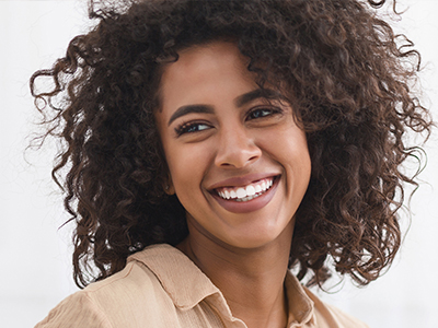 The image displays a smiling woman with curly hair, wearing a beige top, against a light background.